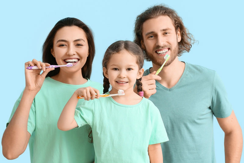 Smiling family of three—mother, father, and young daughter—brushing their teeth together in matching mint green shirts against a light blue background.