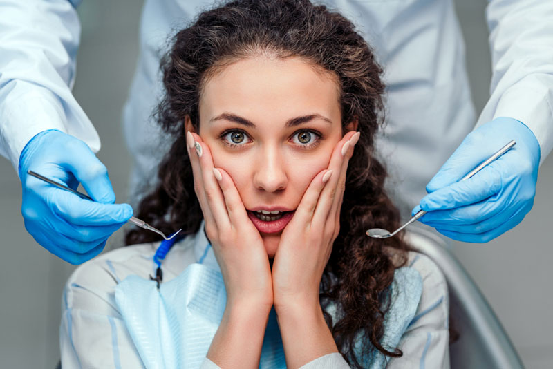 Young woman in a dental chair looking anxious with hands on her face, surrounded by gloved dentists holding dental tools.