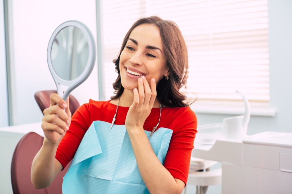 Woman smiling as she looks at her teeth in a hand mirror at a dental office